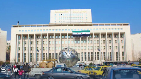واجهة مبنى حكومي في دمشق مع حركة مرور مزدحمة ومجسم كروي معدني وعلم الثورة السورية مرفوع على المبنى، في مشهد يعكس واقع سوريا بين القانون والثقة وصراع مؤسسات الدولة مع تطلعات الشارع.